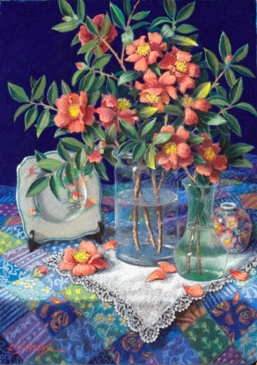 Pink camellias in a glass vase stand on an embroidered cloth. 