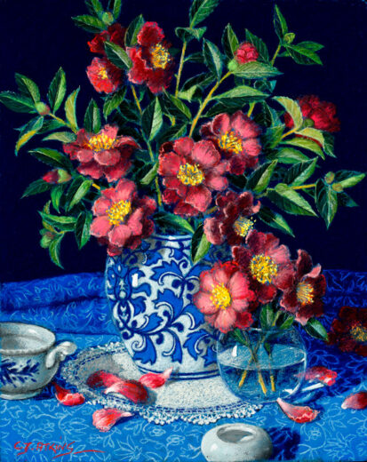 A vase of camellia flowers stand on an lace cloth which itself stands on a patterned blue cloth.