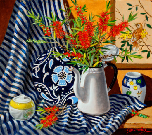 A white enamel coffee pot filled with bottlebrush flowers is in front of a wall hanging featuring a bird.