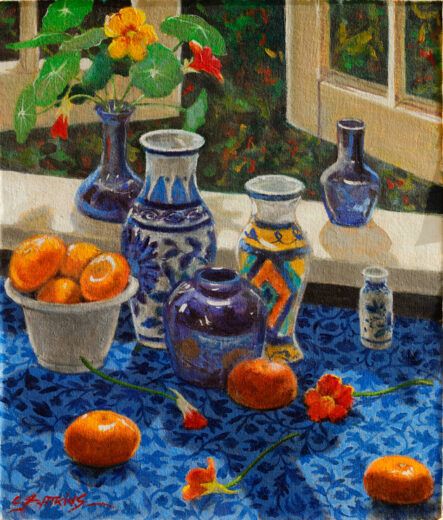 Bright orange coloured fruit and several other objects stand on a patterned blue cloth in front of an open window.