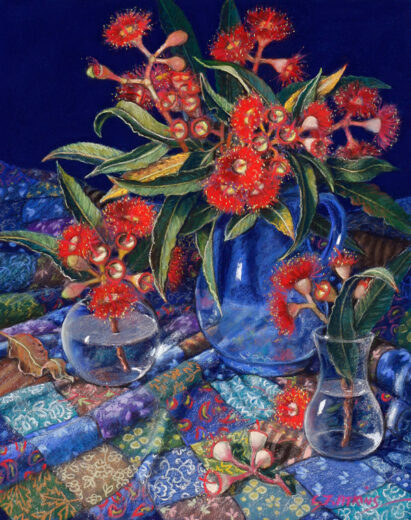 A blue jug and two glass vases contain red flowers. A highly -decorated patchwork cloth is under the vases.